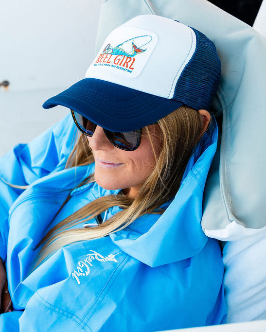 A person wearing a blue jacket and sunglasses, lounging and resting their head on a blue and white trucker hat with a mermaid patch.