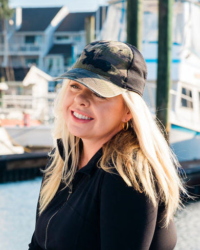 A person wearing a camouflage hat and sunglasses, sitting on the deck of a boat with a marina in the background.
