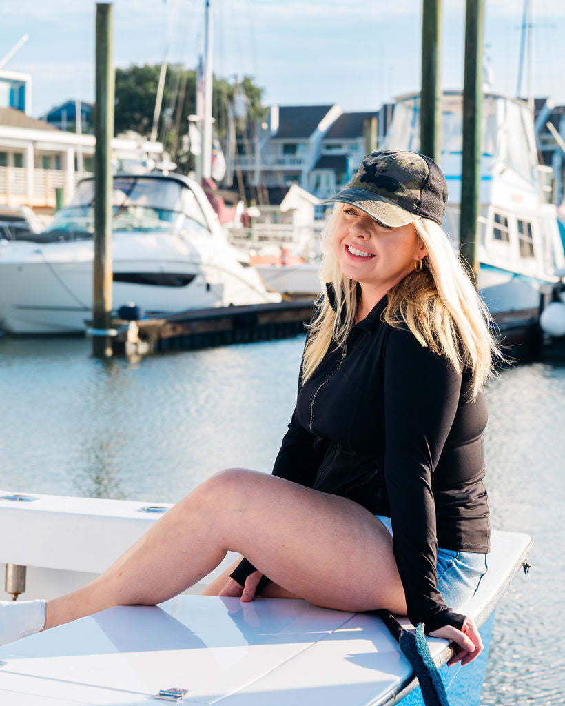 A person wearing a camouflage hat and sunglasses, sitting on the deck of a boat with a marina in the background.